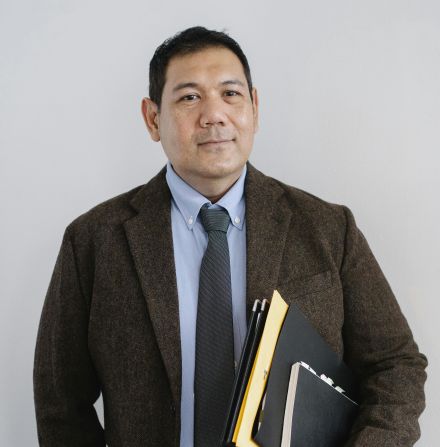 Positive Asian male office worker in formal suit with paper folders in hand standing with hand in pocket against white wall